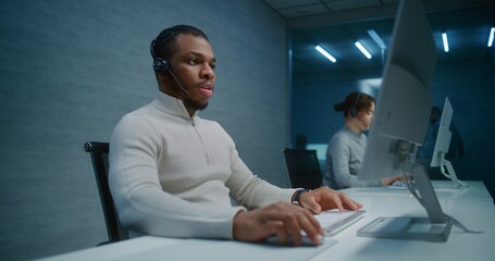 Call center: African American hotline operator talks on headset with customer, works on computer,...