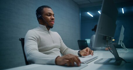 Call center office at night: African American technical support specialist in headset works on PC,...