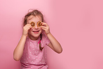 Cheerful young girl holding dried orange slices over her eyes, wearing a heart patterned shirt against a pastel pink background. A fun and playful concept for creative themes.