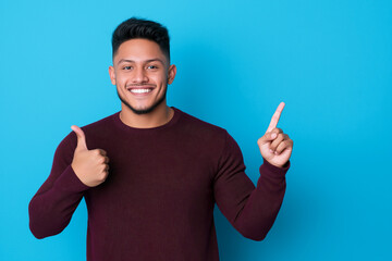 Fototapeta premium young man with cheerful expression is smiling and pointing upwards against blue background, wearing maroon sweater