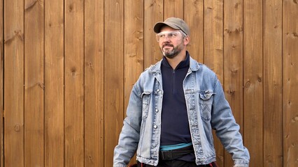 Man standing against wooden wall. Bearded. 40-45 years old. Dressed in denim jacket. Outdoors....