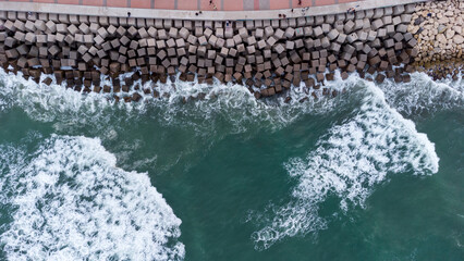 Aerial view of powerful waves crashing against a concrete coastal barrier with adjacent promenade,...