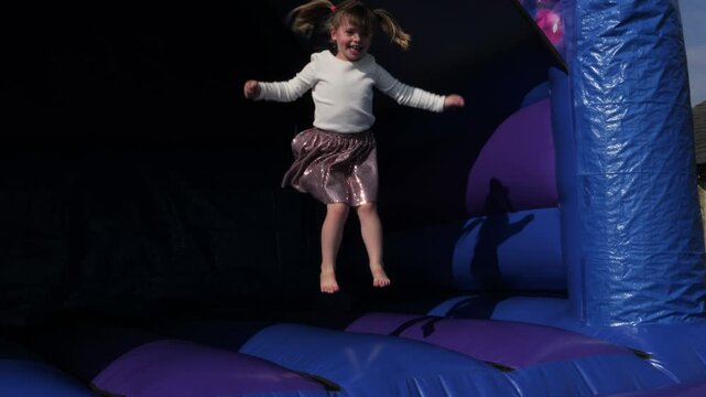4K: Young Girl bouncing  in a party dress on a Bouncy Castle Bounce house Outdoors in summer.  Stock Video Clip Footage