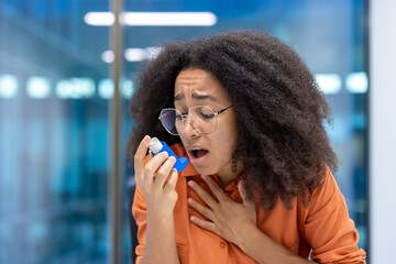 Concerned young woman using asthma inhaler in modern office. She holds her chest, appearing...