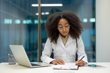 Doctor working diligently in modern office setting, reviewing medical documents with laptop and...