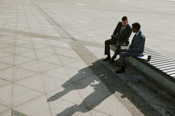 Two businessmen engaged in discussion while sitting on a bench in an outdoor urban area, creating...