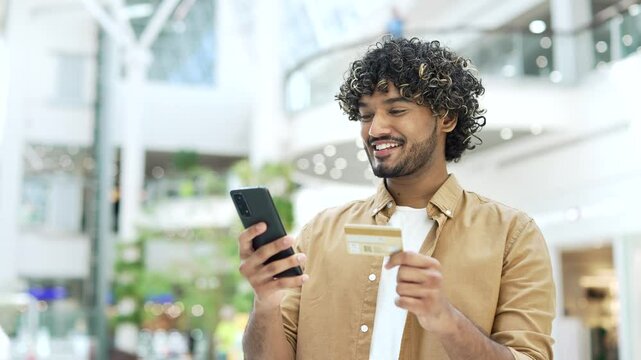 Young handsome man in a dress using smart phone and credit card smiling make online payments standing in a shopping center mall store, Male tourist traveler uses a smartphone doing purchase paid app
