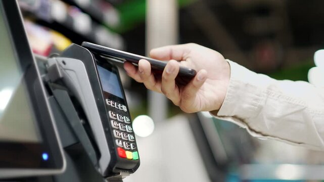 Close-up male hand with a mobile phone in hands pay paid for purchases in stores Contactless NFC Terminal Close up man At Counter Buy electronic payment supermarket smartphone Closeup card reader pos