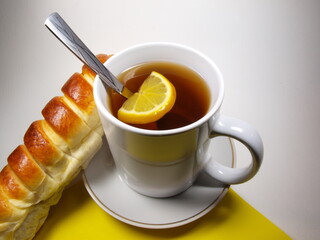 A cup of tea and lemon standing on a saucer. Baked goods - puff pastry, bun. Breakfast