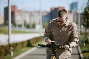 Fototapeta premium Man riding electric scooter on pathway within urban area, focusing on handlebar while background showcases blurred city buildings on sunny day
