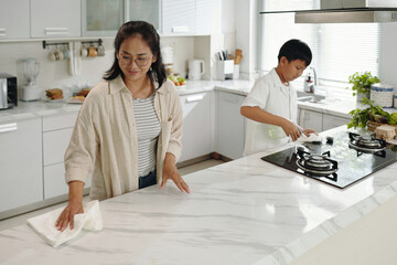 Woman cleaning marble countertop while boy cooking on stove in a bright and airy kitchen featuring...