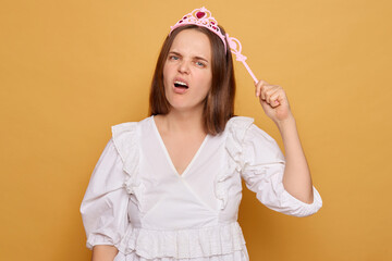 A young woman wearing a white dress and a pink princess crown playfully poses while making a funny...