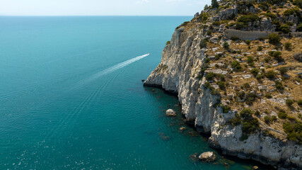 Aerial view of a boat sailing in the Adriatic Sea. On the right is Saraceno Mount. It is a cliff on mediterranean Sea with a trekking path for mountain hiking.