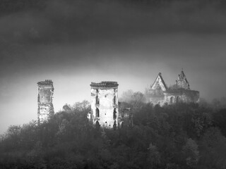 black and white landscape of ruins of medieval castle and church on hill in forest in foggy morning