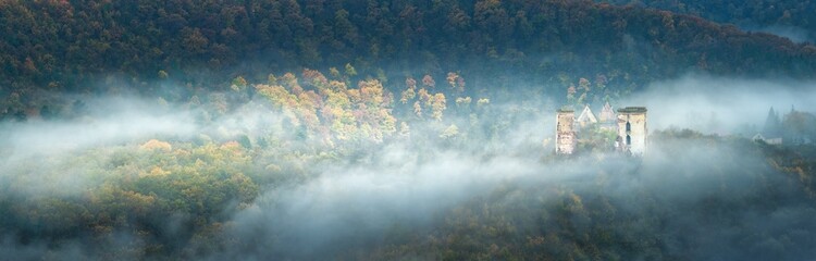 wide-angle panorama of autumn forest with waves of fog and ruins of castle in cold morning