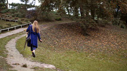 Obraz premium girl walks along the path in the park back view. woman goes into the distance along the autumn alley of the park. a girl in a blue coat is walking on fallen leaves. walk in the fresh air. travel