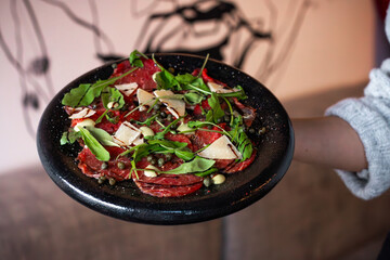 A black plate with beef carpaccio topped with arugula, parmesan shavings, capers