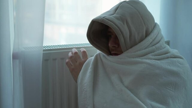 A man in a white blanket sits in a cold apartment.
A man warms himself with a blanket and a warm radiator.
A man in a hood and under a blanket sits next to a warm radiator.
The man is cold