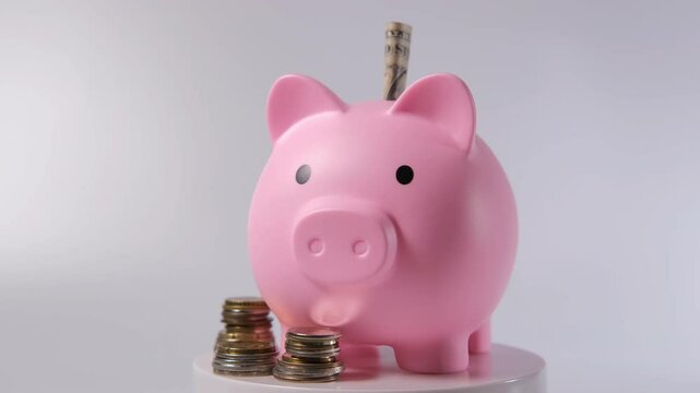Pink piggy bank sits on a rotating pedestal, with coins and banknotes scattered around it by the white background. A banknote peeks out from its back