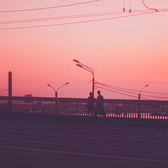 A young couple in love walks across a bridge, with lanterns and wires in the background. The sky is a soft pink, creating a romantic atmosphere.