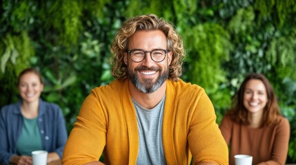Obraz premium cheerful man with curly hair and glasses is positioned at a table in a vibrant cafe surrounded by greenery. Two women are seated in the background, creating a warm and inviting atmosphere