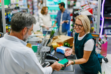 Customer paying with smartphone at supermarket checkout, cashier assisting him