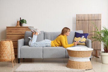Pretty young woman lying on sofa and reading newspaper at home