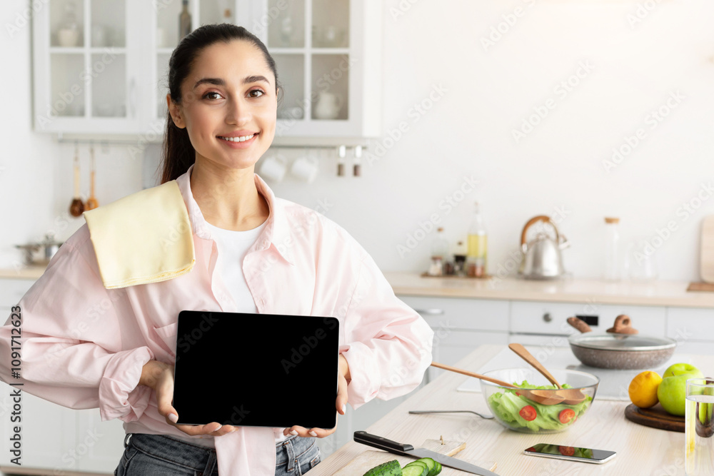 Poster cheerful young lady showing empty digital tablet screen posing standing in modern kitchen at home. s