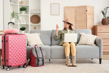 Obraz premium Young woman planning vacation trip online with laptop and luggage at home, travel preparation and booking concept, a pink suitcase and a backpack are placed beside the couch