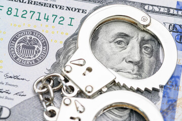 Financial crime, fraud, corruption and legal enforcement : Pair of handcuffs placed on a US dollar 100 bill, visually represents the consequences of illegal financial activities e.g money laundering.