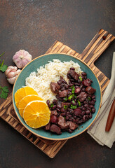 Hearty brazilian feijoada stew with black beans, pork, beef, rice, and orange slices in rustic bowl...