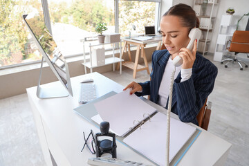 Young businesswoman with folder talking by phone at table in office