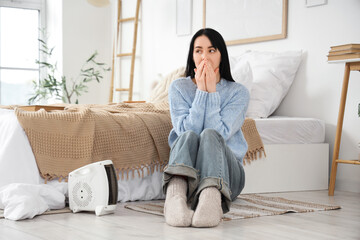 Fototapeta premium Frozen young woman with electric fan heater sitting in bedroom
