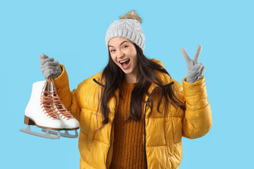 Young woman in winter clothes with ice skates showing victory gesture on blue background