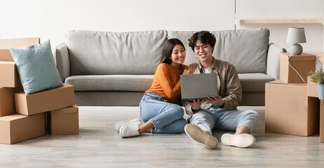 Obraz premium Loving young Asian couple with laptop buying household goods for their new home online, sitting on floor with pc on moving day, copy space. Millennial homeowners planning their house design online