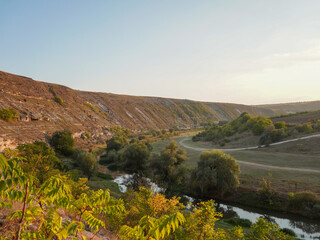 A tranquil river meanders through a parched valley. The sun casts a warm glow on the dry, brown hillsides. Lush green vegetation lines the riverbanks, providing a contrast to the arid landscape.
