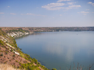 A calm Dniester River stretches under a clear sky. The river curves gently, bordered by a steep, earthy bank covered in sparse vegetation. A small village is visible in the distance across the water.