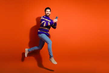 Naklejka premium Full body photo of attractive young man running hold gadget dressed stylish striped purple clothes isolated on orange color background