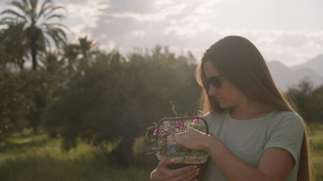 A Woman in Sunglasses Examines a Basket of Easter Eggs and Flowers in a Sunlit Field. Slow motion.