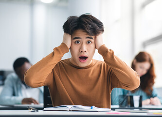 Portrait of shocked asian male student sitting at desk in classroom, grabbing his head looking at camera. Worried youth unprepared for test or exam, thinking about deadline or hard new theme