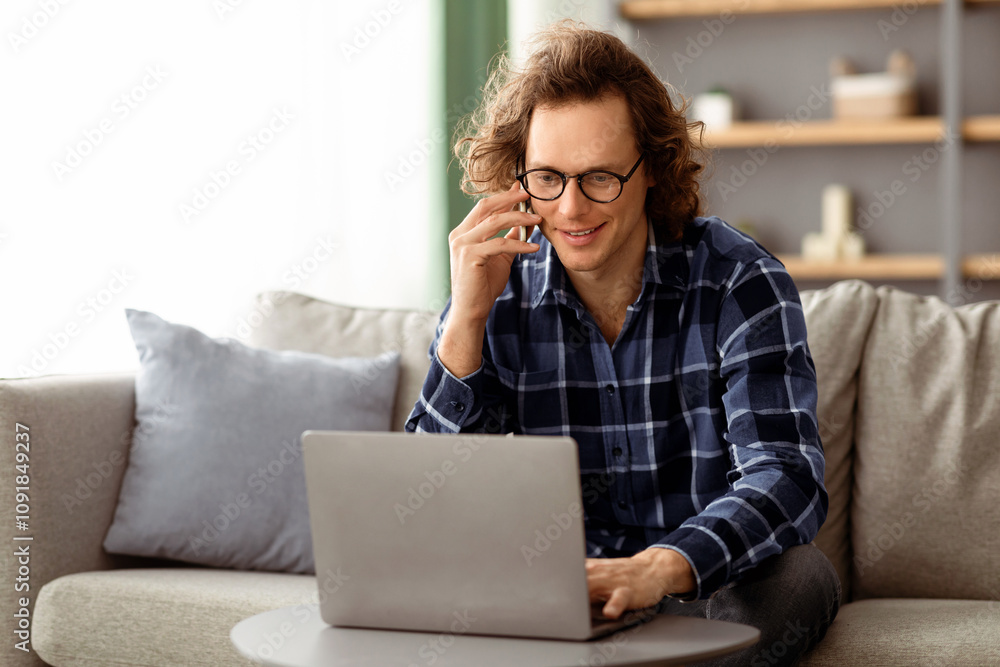 Poster Freelancer Guy Talking On Cellphone Using Laptop Sitting On Sofa Indoor, Working Online Remotely From Home. Hipster Man Having Phone Conversation Communicating And Browsing Internet On Computer