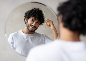 Male haircare. Positive indian bearded guy brushing curly hair with hairbrush, looking and smiling at his reflection in round mirror in bathroom. Hair styling and morning routine concept