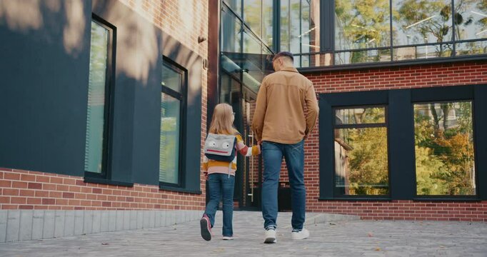 View from behind of Caucasian father and daughter walking together back home from school. Going towards entrance to modern building with big windows. Child carrying backpack. Sunny weather.
