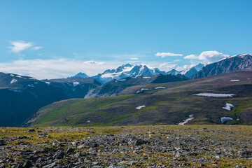 Top view from sunlit grassy stony plain of pass to big snow castle in bright sun under clouds in blue sky. Scenic landscape with green hills and large snow-white rocky mountain far away in sunny day.