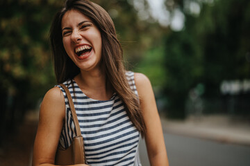 Naklejka premium A joyful young woman with long hair is laughing outdoors, exuding happiness and carefree energy, while wearing a striped dress. The blurred green background enhances the joyful mood of the scene.