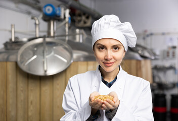 Portrait of positive woman brewmaster holding bunch of malt seeds in beer factory. - Powered by Adobe