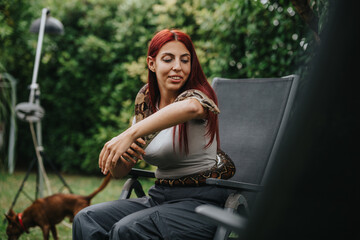 A girl with red hair sits in a garden chair, interacting confidently with a large snake draped over...