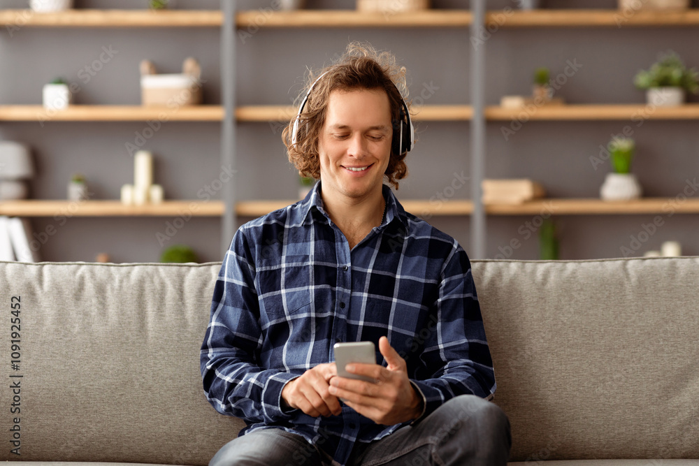 Aufkleber relaxed guy wearing headphones using mobile phone listening to music online sitting on couch at home