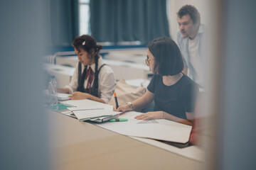 Fototapeta premium Students working diligently in a classroom setting while receiving guidance from a teacher. The focus is on thoughtful expressions and attentive learning environment.