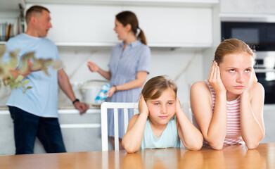 Frightened and frowning children sit at kitchen table during mom and dad quarrel. Behind children, parents swearing in background. Family conflict, divorce